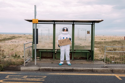 Picture of a spaceman waiting at a bus stop holding a cardboard sign that says Earth